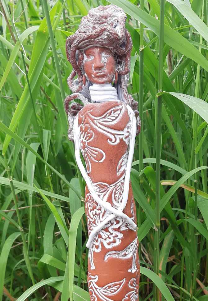 Keramikprodukt von Claudia Entner - Hohe schmale Figur in Terracotta, viele lange, dicke Haare, Körper schlicht und gerade mit gestempeltem Blumenmotiv, dass mit weisser Glasur ausgefüllt ist, sie trägt runde Ohrringe, Halsringe, die Hände sehr dünn und lange kreuzen sich über ihrem Schoßraum