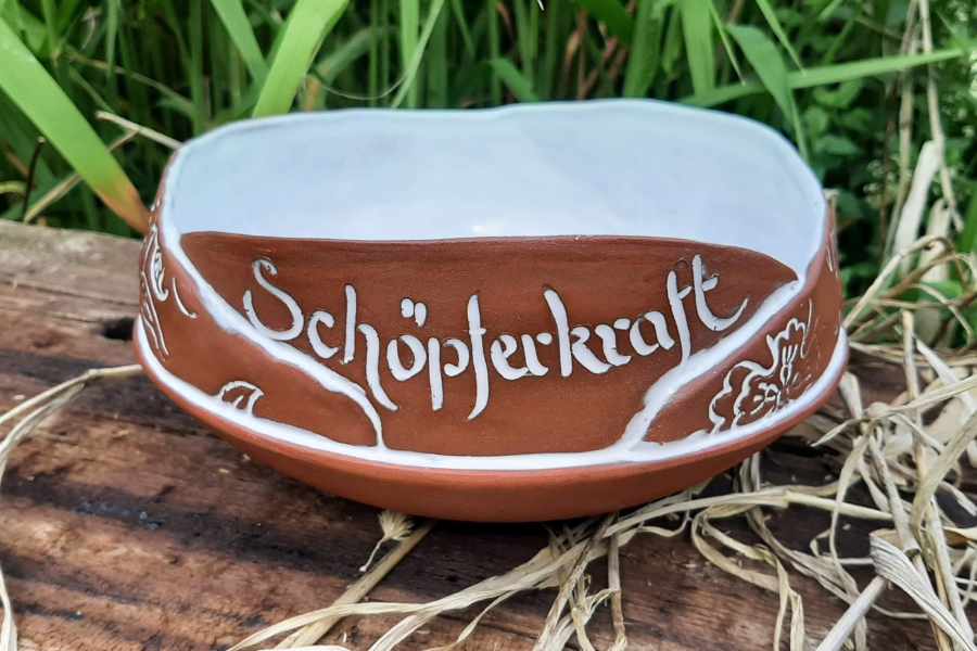 Keramikprodukt von Claudia Entner - Schale in Terracotta-Rot, gerades aufgesetztes Band 3-4cm, das gestempelt ist mit Blumen und Blätterdesign, das wiederum mit weisser Glasur gefüllt ist, in weiss eingelegt der Schriftzug 'Schöpferkraft'.