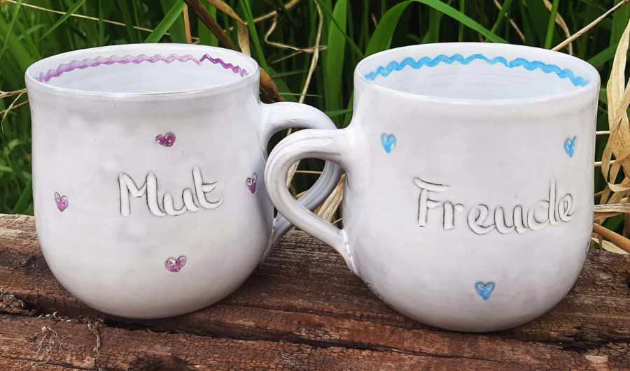 Keramikprodukte von Claudia Entner - 2 bauchige Becher, deren Henkel zueinander gerichtet sind, links lila Herzen Aufschrift 'Mut' rechts blaue Herzen Aufschrift ' Freude'