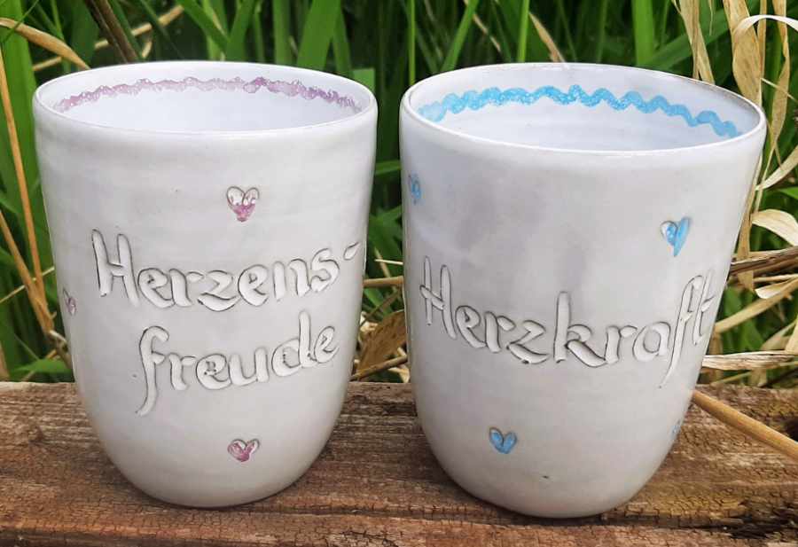 Keramikprodukte von Claudia Entner - 2 Becher weiß glasiert, links lila Herzen Aufschrift 'Herzensfreude', rechts blaue Herzen Aufschrift 'Herzkraft'