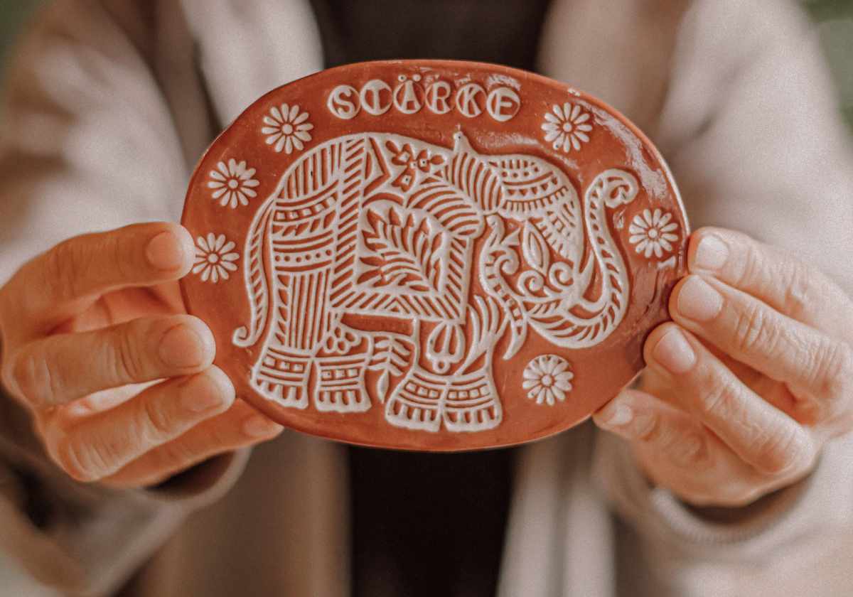 Claudia Entner Keramikprodukte: Hände die nach vorne eine terracottarote Tontafel halten mit dem Motiv eines Elefanten eingestempelt, mit der Aufschrift "Stärke"